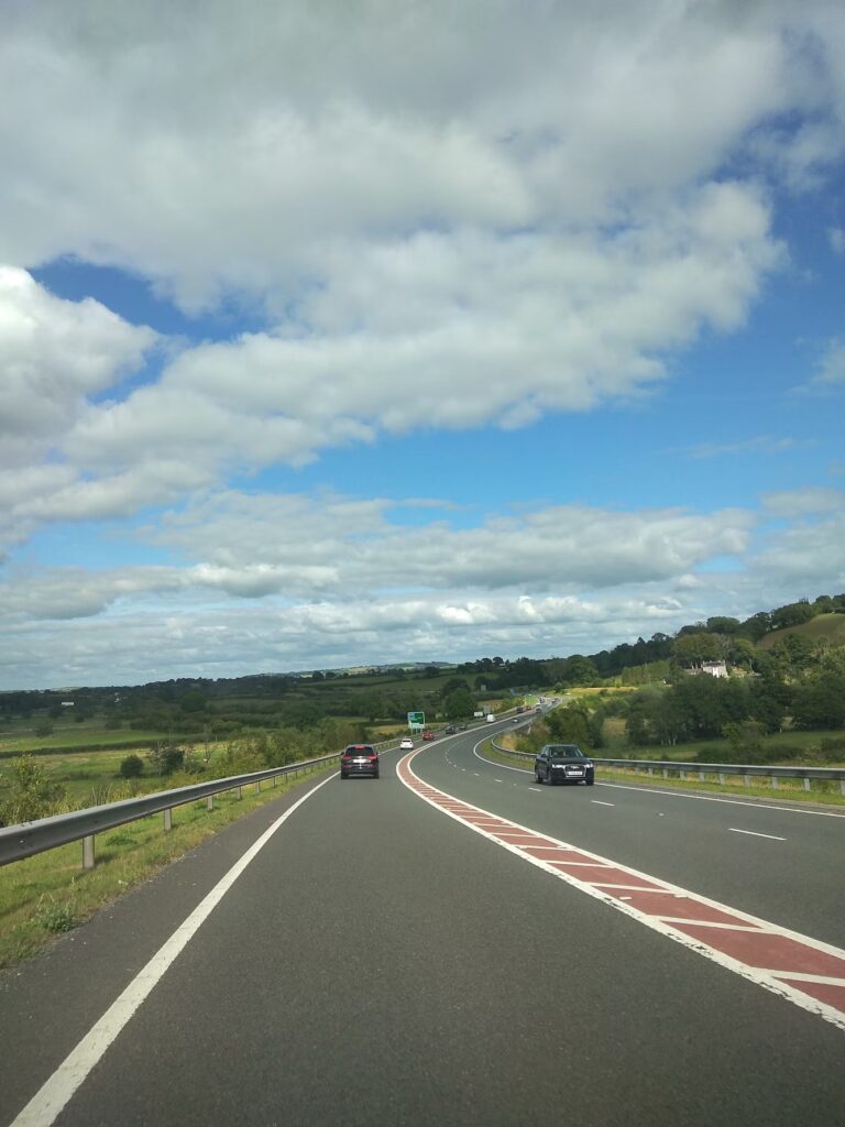 A road through the countryside with several cars driving in both directions. A blue sky with several clouds